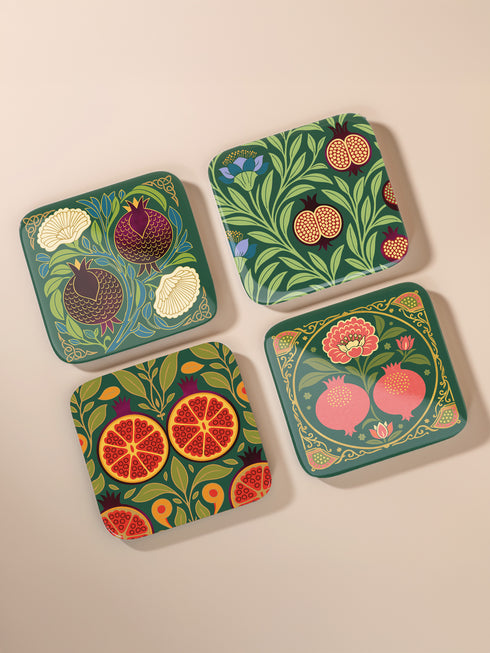 Set of four square drinking coasters featuring rich, dark green botanical illustration with highly detailed Pomegranate fruits and sliced cross-sections. The ornate pattern utilizes deep crimson, opulent gold, and ivory tones, strongly reflecting the stylized Art Nouveau period aesthetic and William Morris textile designs. Crafted from durable, resilient masonite hardboard, this luxury item features a protective high-gloss finish and a non-slip cork base.